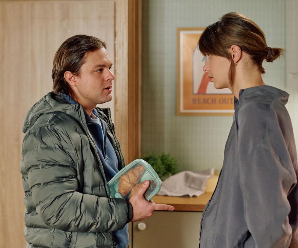 Mężczyzna w ciemnej kurtce puchowej i kobieta w szarej bluzie rozmawiają w kuchni. On trzyma pudełko z rogalikami, ona słucha z poważną miną. Scena z serialu Na Wspólnej, gdzie Remigiusz dowiaduje się o umierającym ojcu. Więcej o wydarzeniach z ul. Wspólnej na Super Seriale.