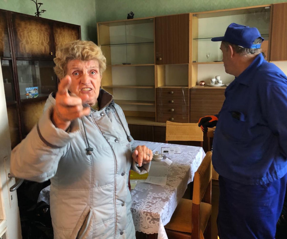 Zagniewana starsza kobieta wskazuje palcem na mężczyznę w niebieskim ubraniu roboczym i czapce, stojącego tyłem w mieszkaniu. W tle meblościanka i stół z obrusem. Zdjęcie ilustruje temat samowolnej eksmisji lokatora na Super Biznes.