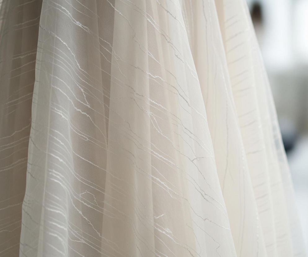 Pofałdowany, kremowy materiał wypełnia większą część kadru, tworząc miękkie, pionowe fałdy. Na jego powierzchni widoczne są nieregularne, cienkie, srebrzyste linie przypominające wzór marmuru lub pęknięcia. Tkanina jest półprzezroczysta, delikatnie przepuszcza światło. Tło jest rozmyte, ukazując jasne, niewyraźne kształty.