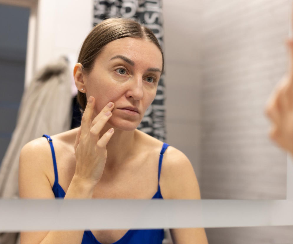 Kobieta z ręką dotykającą policzka, spoglądająca w lustro z zaniepokojeniem. Ilustracja symbolizuje problem zmian rysów twarzy, o którym przeczytasz więcej na Poradnik Zdrowie. Twarz kobiety ma neutralny wyraz, a jej włosy są związane w niski kucyk.