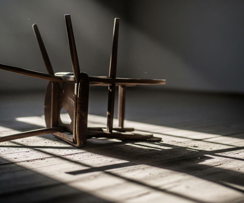 Na drewnianej podłodze, pokrytej kurzem i drobnymi cząsteczkami, leży przewrócone drewniane krzesło. Krzesło jest brązowe, jego nogi i oparcie skierowane są w górę, a siedzisko leży płasko na deskach. Światło słoneczne wpada przez niewidoczne okno po lewej stronie kadru, rzucając długie, prostokątne cienie na podłogę i oświetlając kurz w powietrzu. W tle widać fragment białego okna i ciemną ścianę z niewyraźnym zarysem grzejnika.