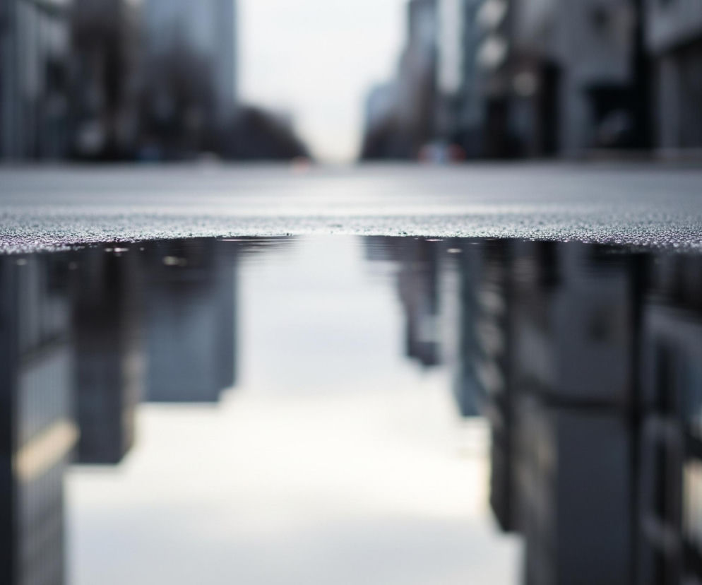 Na pierwszym planie widoczna jest mokra, ciemnoszara nawierzchnia drogi z licznymi drobnymi kropelkami wody. Pośrodku kadru znajduje się kałuża, w której odbija się otoczenie. Odbicie ukazuje rozmyte kształty wysokich, ciemnych budynków, przypominających miejski krajobraz. Powyżej kałuży, w tle, widoczne są te same budynki, jednak również rozmazane, tworząc wrażenie głębi i perspektywy z niskiego punktu widzenia. Całość utrzymana jest w chłodnej palecie barw, dominują szarości i ciemne odcienie niebieskiego, z jasnym, zamglonym niebem w oddali.