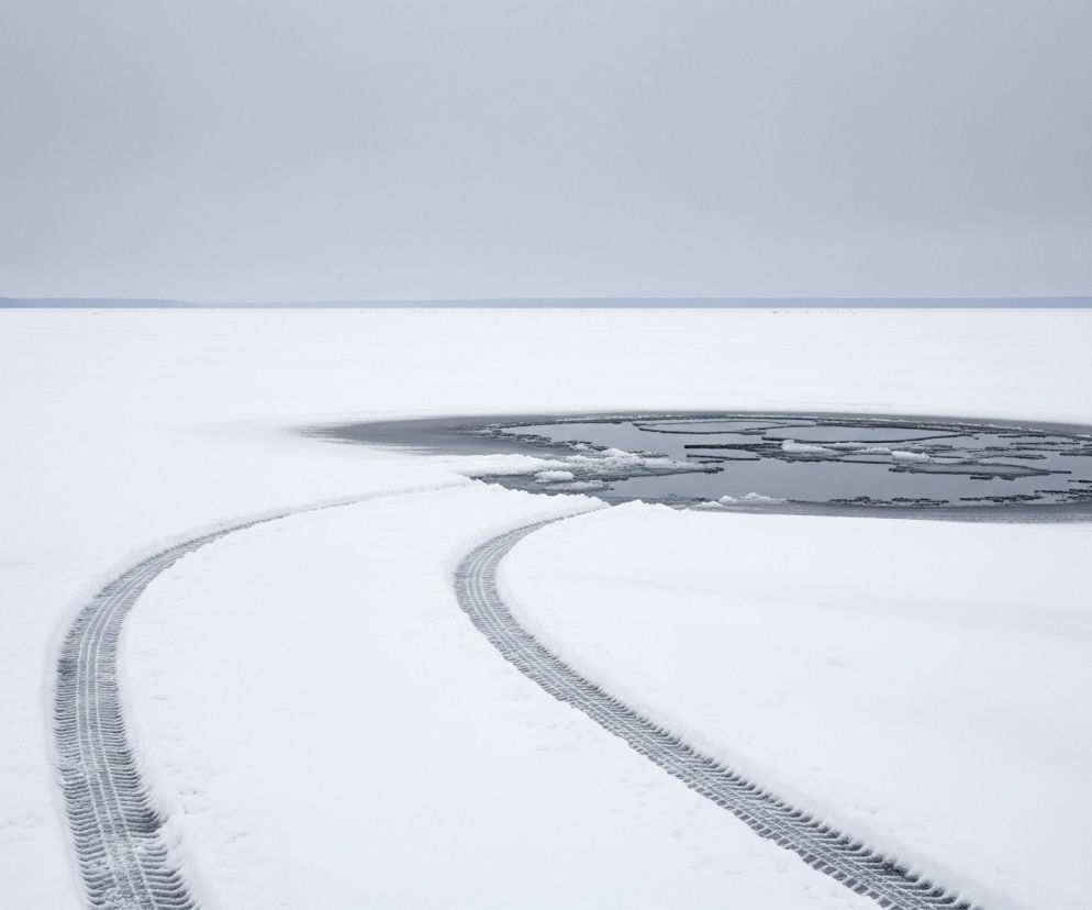 Zamarznięta, zaśnieżona powierzchnia dominująca w dolnej części obrazu ciągnie się w stronę horyzontu. W prawym górnym kwadrancie, na tej białej tafli, widoczny jest okrągły przerębel z ciemną wodą, w której pływają fragmenty lodu. Od lewego dolnego rogu rozchodzą się dwa równoległe ślady opon, wygięte łukowato w prawo, prowadzące do krawędzi przerębla. Horyzont tworzy cienka, ciemniejsza linia, nad którą rozpościera się jednolite, jasnoszare niebo.