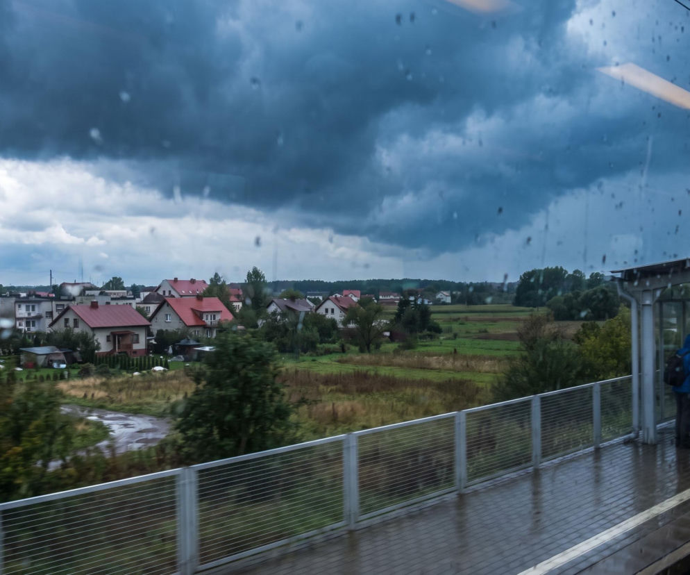 Ciemne chmury burzowe nad osiedlem domów widziane z mokrego okna pociągu. O pogodzie w Polsce przeczytasz na Eska.
