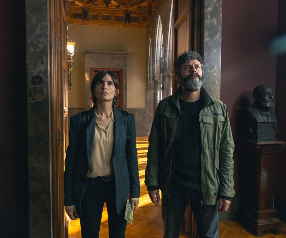 Dwoje ludzi, detektywów z serialu Miasto Cieni na Netfliksie, w eleganckim wnętrzu budynku autorstwa Antonio Gaudiego, patrzą w górę. Oboje są wyraźnie zamyśleni i skupieni na czymś poza kadrem. Po prawej stronie widać postument z ciemnym popiersiem. Więcej o Gaudim na Architektura Murator Plus.