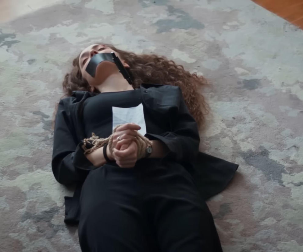 Nana leżąca na wzorzystym dywanie, ubrana na czarno, z ustami zaklejonymi czarną taśmą i rękami związanymi sznurem, obok niej biała kartka. Scena grozy z serialu Dziedzictwo, o której przeczytasz więcej na Super Seriale.