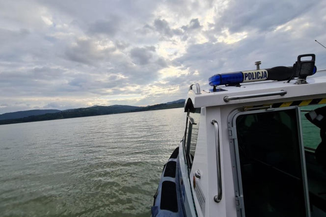 Policyjna łódź patrolowa na wodach jeziora, z widocznymi niebieskimi sygnałami świetlnymi i napisem POLICJA. W tle rozciąga się linia brzegowa zalesionych wzgórz pod zachmurzonym niebem. Więcej o bezpieczeństwie nad wodą przeczytasz na naszym portalu.