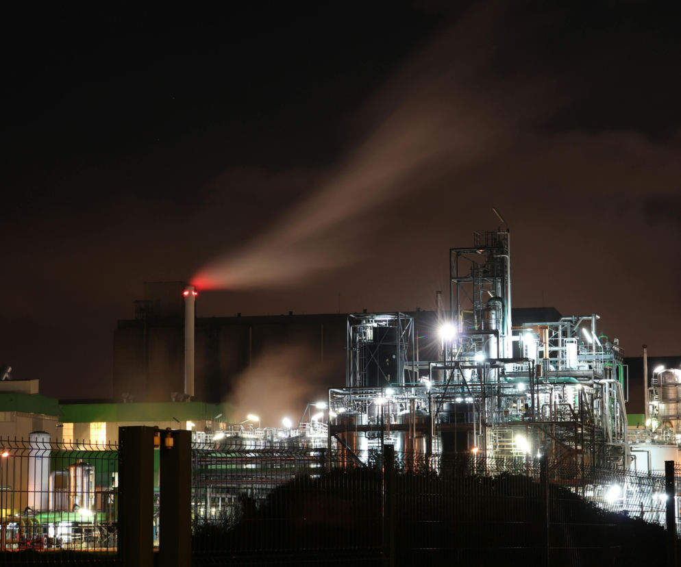 Nocny widok na fabrykę chemiczną, z której komina wydobywa się dym podświetlony na czerwono, symbolizuje kryzys przemysłowy w Europie. Instalacje przemysłowe oświetlone licznymi lampami stoją na tle ciemnego nieba. Więcej na Super Biznes.