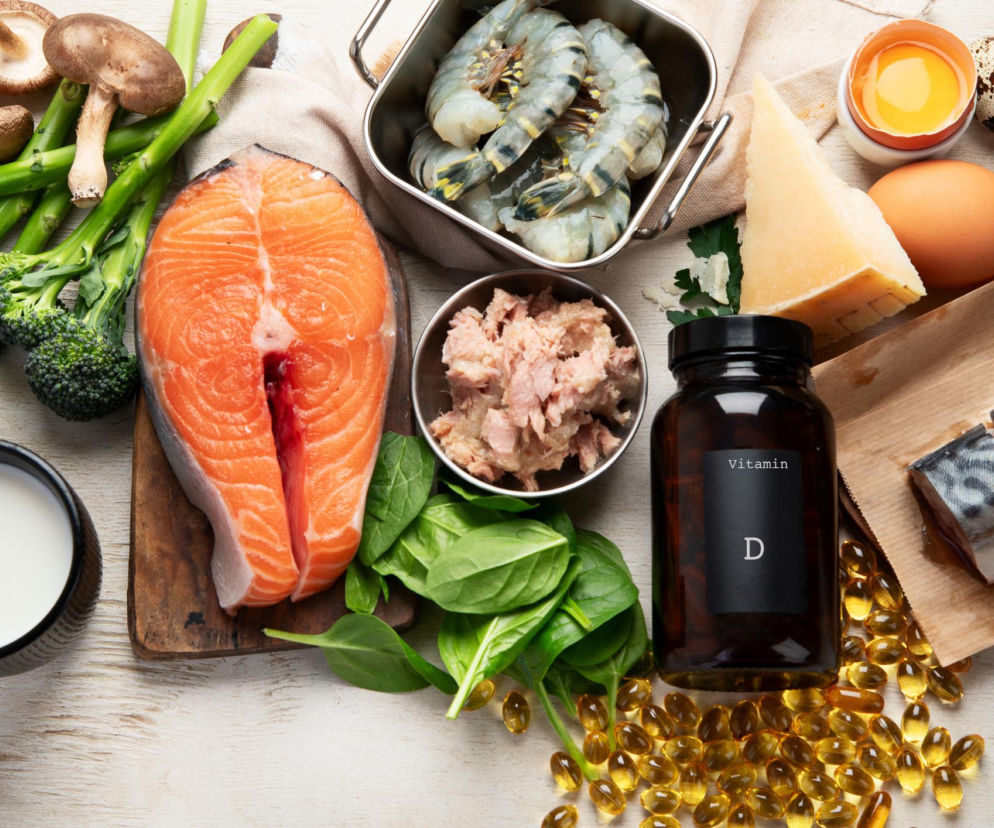 Rozłożone na stole produkty bogate w witaminę D: świeży łosoś, makrela, krewetki, tuńczyk, jajka, mleko, sery, pieczarki, brokuły, szpinak, obok nich buteleczka z napisem „Vitamin D” oraz żółte kapsułki suplementu. Więcej o witaminie D i jej wpływie na zdrowie przeczytasz na Poradnik Zdrowie.
