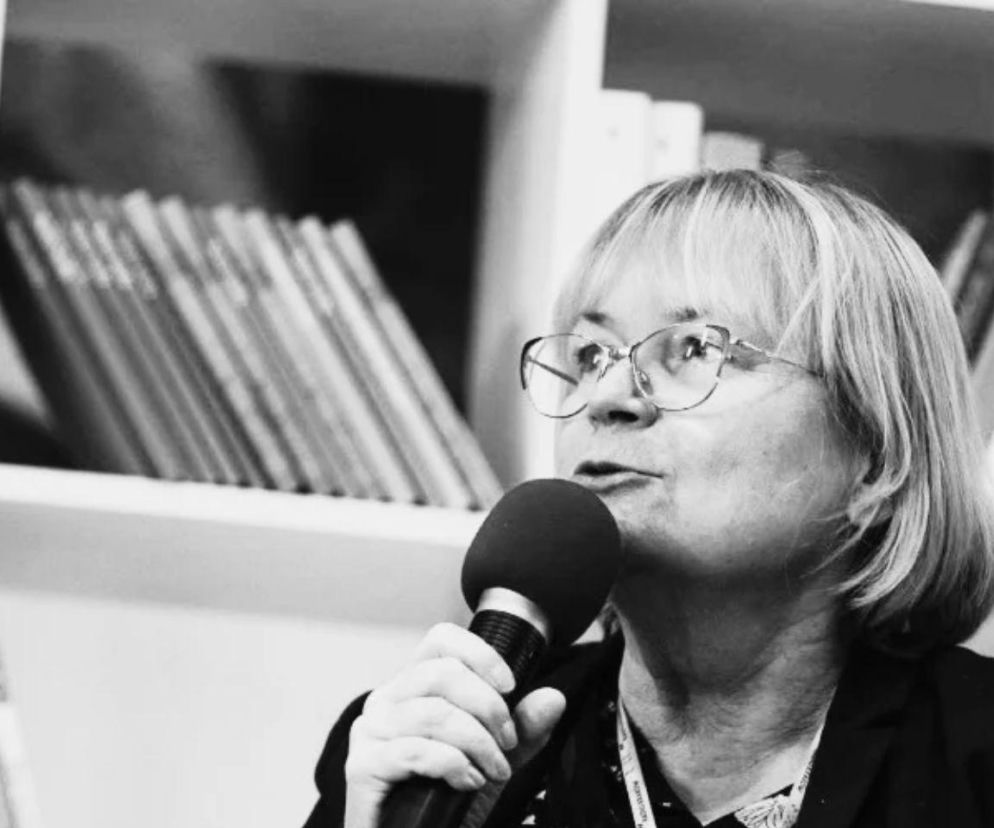 Barbara Gawryluk w okularach przemawia do mikrofonu na tle regału z książkami. O śmierci pisarki przeczytasz na SE.