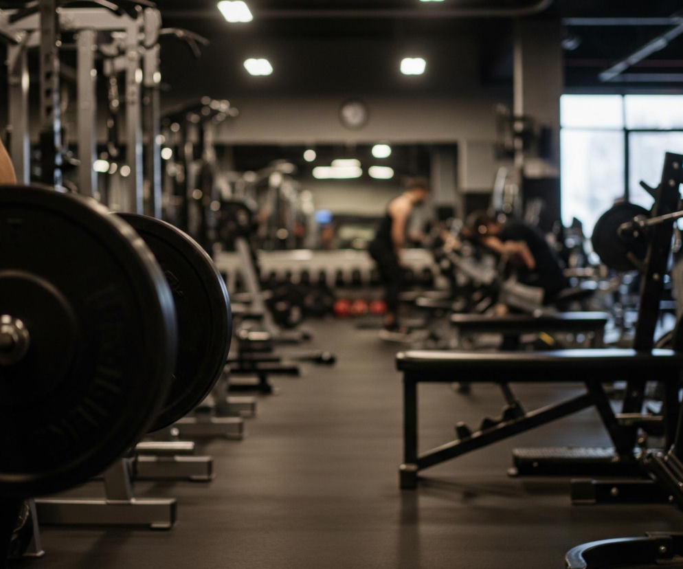 Na pierwszym planie, po lewej stronie kadru, widoczny jest tors osoby w czerwonej koszulce, trzymającej w rękach sztangę z dużymi, ciemnymi talerzami. Podłoga w siłowni jest ciemnoszara, a sprzęt, w tym ławki i stojaki na ciężary, utrzymany jest w czarnej i srebrnej kolorystyce. W tle widać niewyraźne sylwetki innych ludzi ćwiczących na różnych maszynach oraz duże okno, przez które wpada światło. Całe pomieszczenie jest oświetlone ciepłym, rozproszonym światłem z górnych lamp.