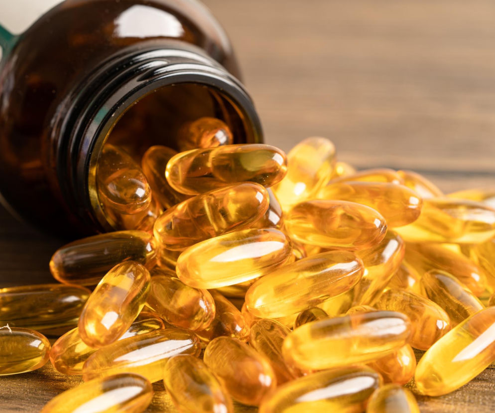 Żółte, przezroczyste kapsułki z olejem rybim, bogate w kwasy omega-3, rozsypane obok szklanego brązowego słoiczka na drewnianym tle, symbolizujące suplementację korzystną dla zdrowia. Dowiedz się więcej o ich wpływie na cukrzycę typu 2 na PoradnikZdrowie.