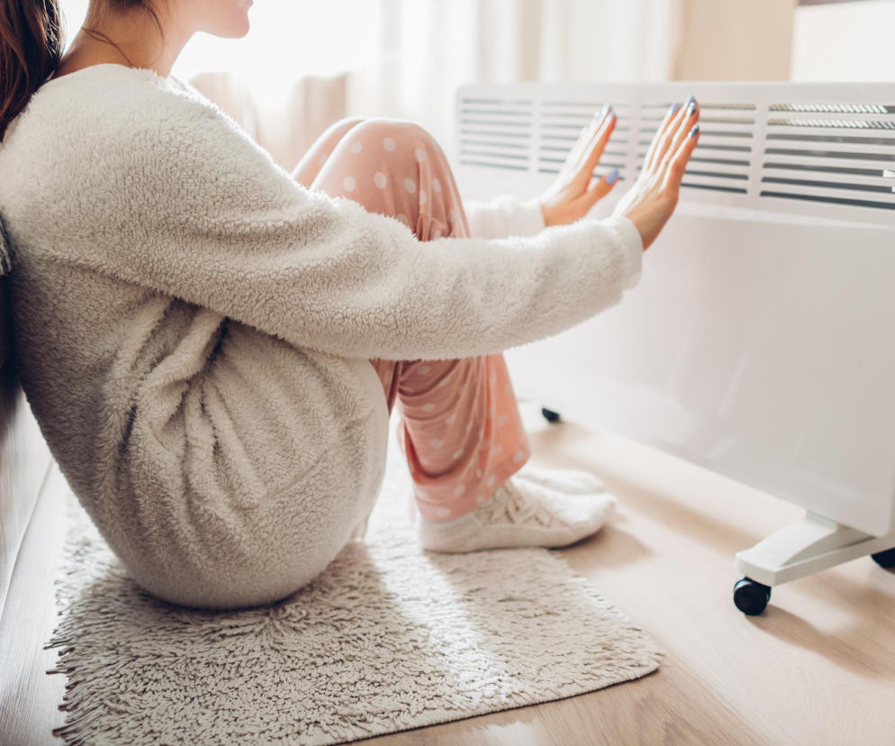 Kobieta w puszystym, beżowym dresie i różowych spodniach z białymi kropkami ogrzewa dłonie przy białym grzejniku konwektorowym, siedząc na drewnianej podłodze, co ilustruje problem zimnych dłoni i stóp, o którym można przeczytać na Poradnik Zdrowie.