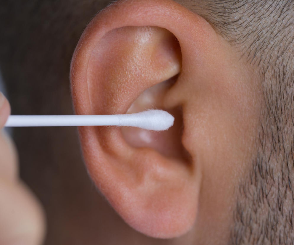 Ucho dorosłego mężczyzny czyszczone patyczkiem higienicznym, co może wywoływać kaszel, zgodnie z artykułem Poradnik Zdrowie o odruchu Arnolda. Mężczyzna ma krótkie, ciemne włosy na skroni.