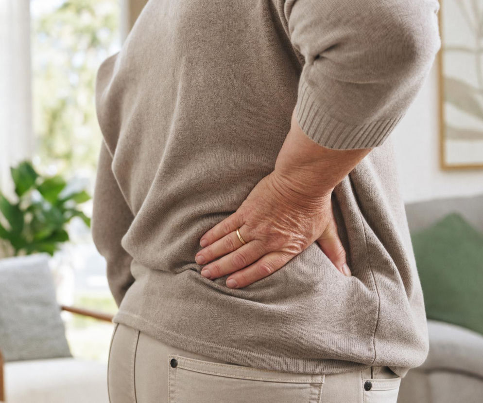 Dłoń starszej osoby uciskająca bolące plecy na wysokości lędźwi. O objawach osteoporozy przeczytasz na Poradnik Zdrowie.