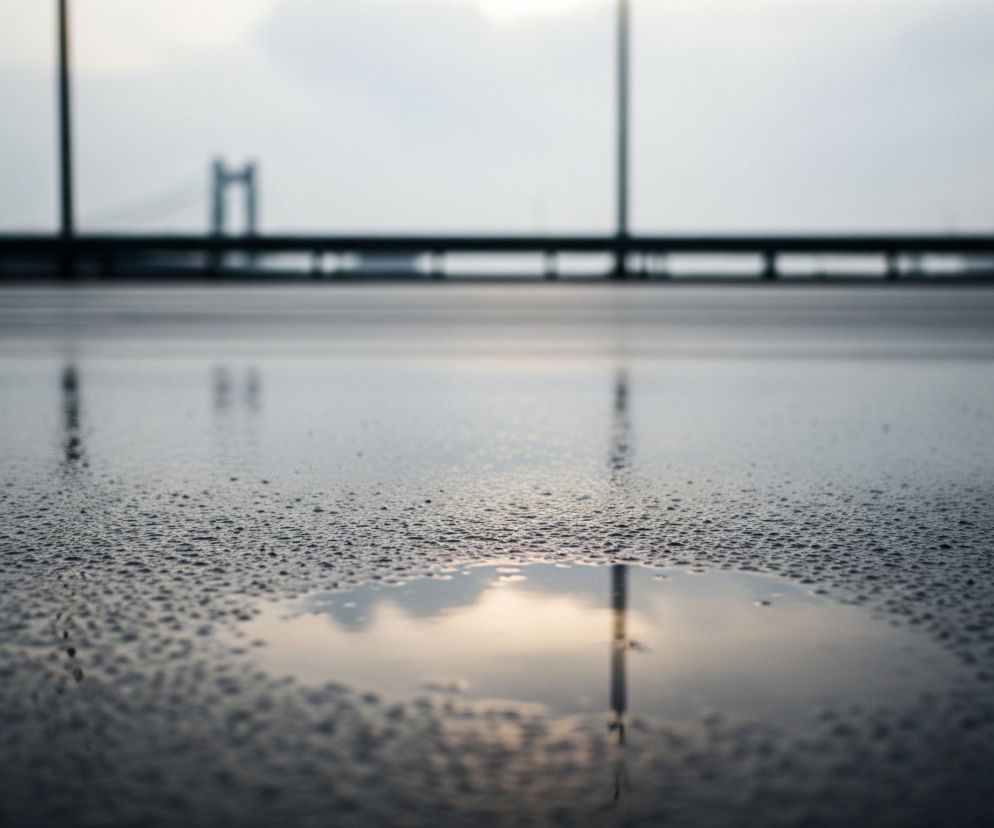 Szara, mokra powierzchnia pokryta jest drobnymi kropelkami wody, co sugeruje niedawny deszcz. W centralnej części pierwszego planu widoczna jest kałuża, w której odbija się jasne niebo z delikatnymi obłokami i element pionowej struktury, prawdopodobnie słupa lub latarni. Na horyzoncie, rozciągającym się wzdłuż górnej części kadru, rysuje się ciemny, rozmyty kontur mostu z dwoma charakterystycznymi wieżami, a także pionowe słupy i poziome belki balustrady lub barierki. Całość utrzymana jest w chłodnej palecie barw, z dominującymi szarościami i błękitami, przełamanymi jedynie ciepłym, żółtawym odbiciem światła w kałuży.