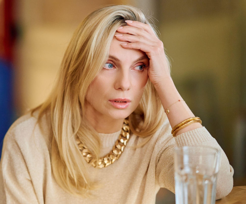 Zmartwiona kobieta z blond włosami, niebieskimi oczami, w beżowym swetrze i złotym naszyjniku, opiera dłoń na czole, spoglądając w bok, obok szklanki wody. Scena oddaje napięcie i trudne emocje bohaterów serialu „Na Wspólnej”, o których można przeczytać na portalu Super Seriale.