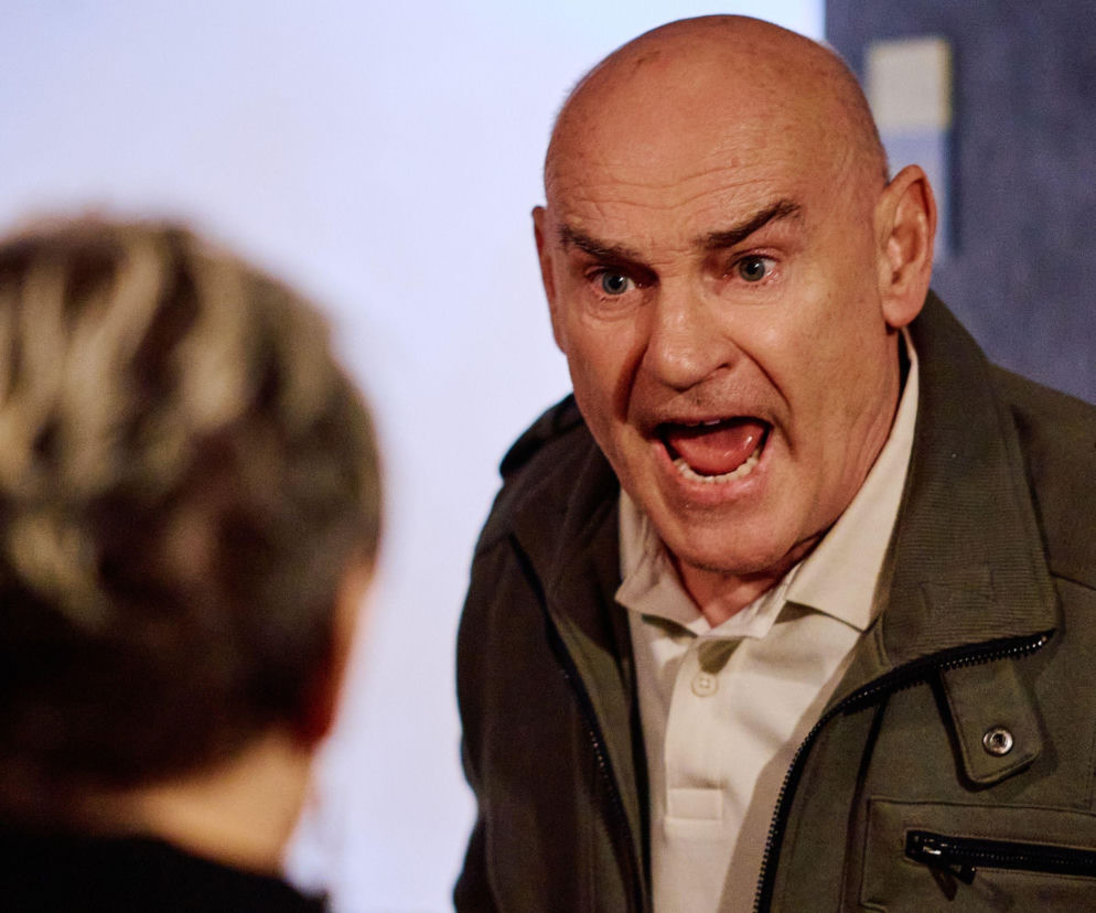 Roman Hoffer z otwartymi ustami, wyrażający szok lub krzyk, ubrany w zieloną kurtkę i jasną koszulkę polo, na tle niewyraźnej sylwetki innej osoby. To emocjonalna scena z serialu Na Wspólnej, o której więcej dowiesz się na Super Seriale.