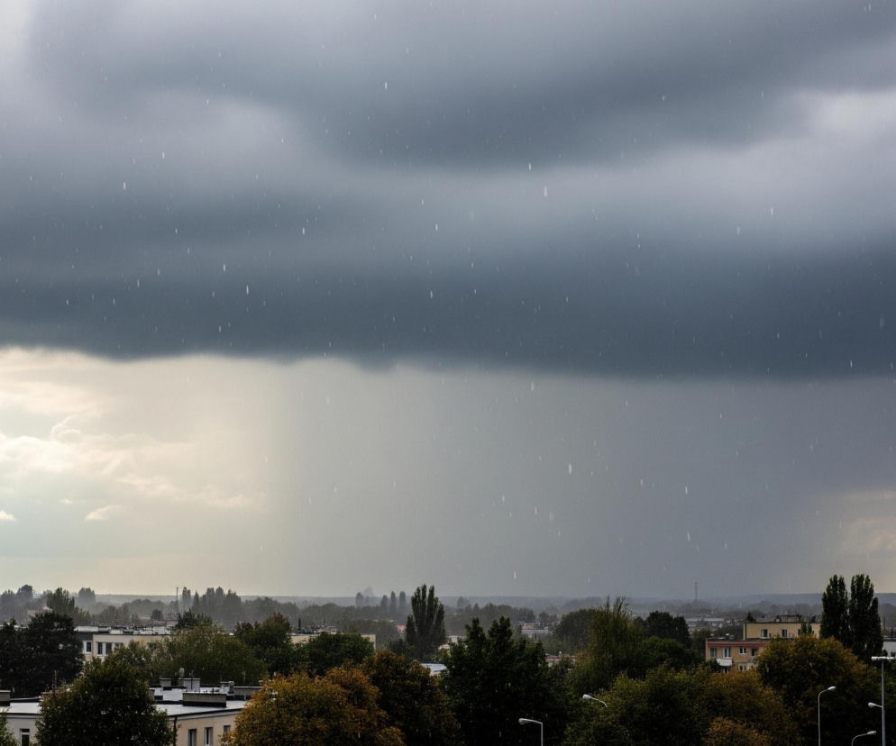 Kadr przedstawia miejski pejzaż podczas deszczu, z dominującym ciemnoszarym niebem i widocznymi, pionowymi smugami spadających kropel deszczu. W górnej części kadru ciężkie, ciemne chmury rozciągają się horyzontalnie, przechodząc w jaśniejszy, mleczno-szary odcień w kierunku horyzontu. Poniżej rozciąga się pasmo zielonych drzew w różnych odcieniach, z częściowo widocznymi dachami budynków mieszkalnych o białych i beżowych elewacjach, a także latarniami ulicznymi w dolnej części kadru. W oddali, na horyzoncie, rysują się niewyraźne sylwetki wysokich kominów i wież, zanurzone w szarej mgle.