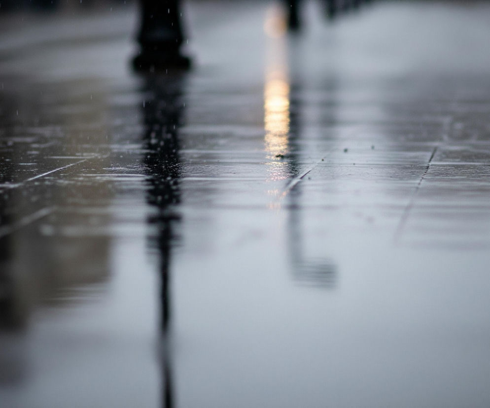 Mokra, lśniąca powierzchnia chodnika, podzielona na prostokątne płyty, dominuje w kadrze, odbijając otoczenie. W centralnej części obrazu widoczne są rozmyte odbicia słupów i niewyraźnych sylwetek, z jednym jaskrawym, pionowym smugą światła w kolorze żółto-pomarańczowym. Na całej mokrej powierzchni rozrzucone są drobne krople wody, tworzące delikatne wzory na szarej nawierzchni. Górna część obrazu, poza pasem odbić, jest bardzo rozmyta i ciemna, przedstawiając nieostre kształty, prawdopodobnie ludzi lub drzew.