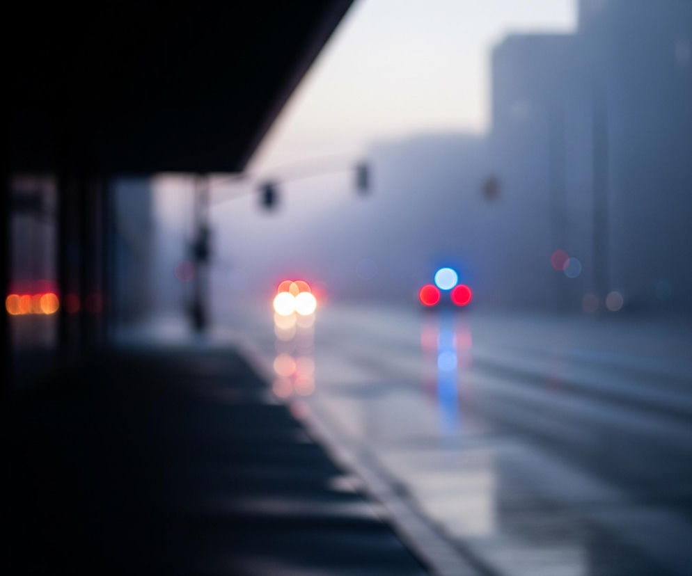 Ujęcie z perspektywy ulicy wieczorem lub wcześnie rano, w pochmurny dzień, charakteryzuje się efektem rozmycia. Od lewej strony obrazu, w dolnym narożniku, znajduje się ciemny, niemal czarny obszar chodnika, rozciągający się w głąb kadru i lekko w prawo, oddzielony od jezdni białą linią. Na drodze widać odbicia świateł pojazdów w kałużach, z dominującymi czerwonymi i pomarańczowymi światłami tylnymi oraz pojedynczym, jasnym niebieskim światłem z przodu, prawdopodobnie samochodu służbowego. Tło jest zamglone, zarysowują się na nim niewyraźne kształty budynków i sygnalizacji świetlnej.