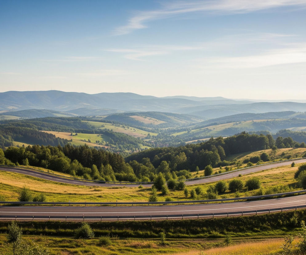 Kręta, asfaltowa droga z białymi liniami biegnie przez pagórkowaty krajobraz, wijąc się przez zielone i żółte pola. Na drodze, w oddali po prawej stronie, jedzie ciemny samochód osobowy. Krajobraz jest porośnięty drzewami i krzewami, a w tle widać liczne pasma gór, których intensywność barw maleje wraz z oddalaniem się, przechodząc od ciemnozielonych do błękitnych odcieni. Błękitne niebo z kilkoma delikatnymi, białymi chmurami rozciąga się nad górami.