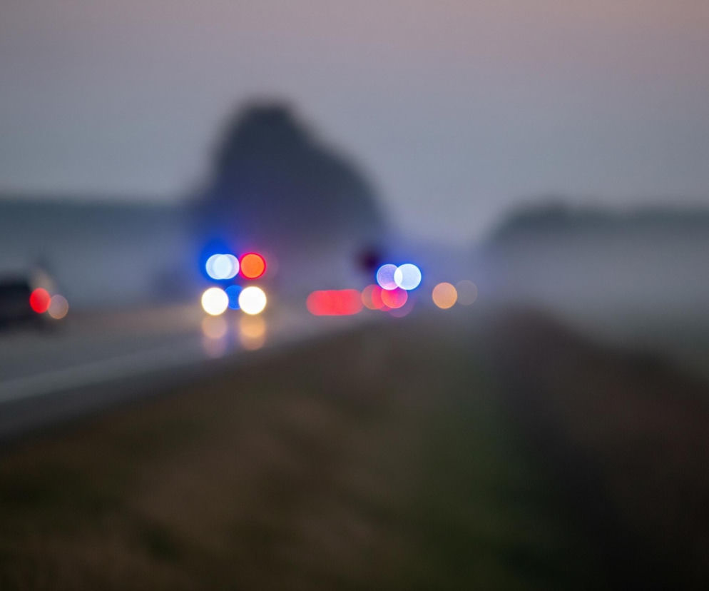 Rozmyty obraz przedstawia drogę o zmierzchu lub w nocy, z niewyraźnymi światłami pojazdów i otoczenia. Na pierwszym planie, po lewej stronie, widoczne są światła tylne czerwonego samochodu, który oddala się od widza. W oddali, na środku obrazu, zlewają się w plamy światła pojazdów, z dominującymi niebieskimi i czerwonymi barwami, a także jasnymi żółtymi lub białymi punktami. Droga ma jasne, odbijające światło linie rozdzielające pasy, a po prawej stronie widać rozmazaną, ciemną linię pobocza lub zarośli. Tło to rozmyte, ciemne sylwetki wzniesień lub drzew, a niebo ma szarofioletowy odcień.