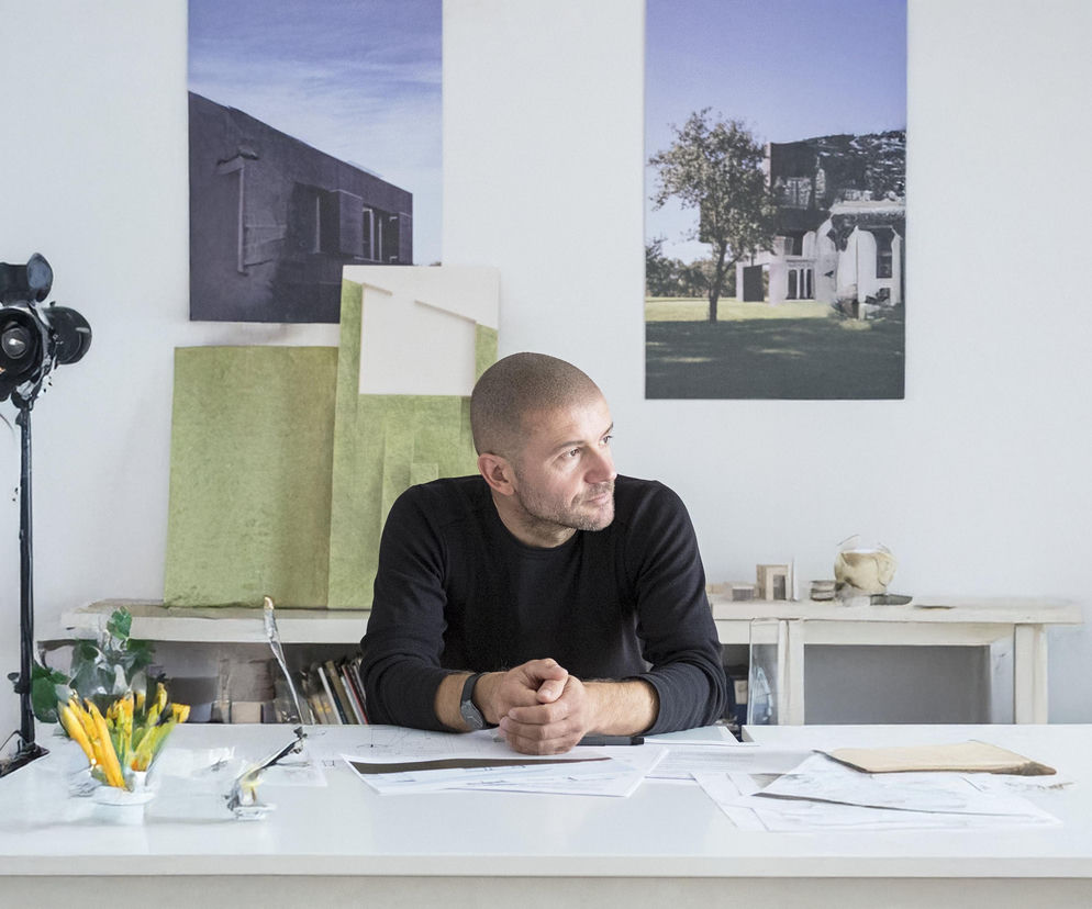 Architekt Robert Konieczny, ubrany w czarny sweter, siedzi przy białym biurku z dokumentami i rysunkami. Jego spojrzenie skierowane jest w bok, symbolizując refleksję nad etycznym projektowaniem. W tle widać dwie abstrakcyjne prace i sprzęt fotograficzny, tworząc profesjonalne otoczenie. O zmianach w myśleniu o architekturze przeczytasz na Architektura Murator Plus.