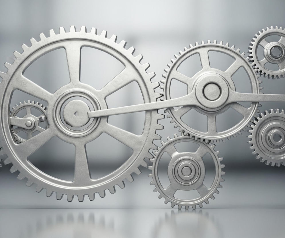 Na obrazie widnieje skomplikowany mechanizm składający się z dziewięciu metalowych kół zębatych, wszystkie w odcieniach srebra i szarości. Duże koło zębate w centrum lewej strony obrazu jest połączone z mniejszymi kołami wokół niego oraz długim ramieniem, które łączy je z innymi kołami po prawej stronie. Koła są różnej wielkości, z zębami na obwodzie, i mają szprychowe konstrukcje wewnątrz, a także widoczne są osie obrotu. Cały system znajduje się na gładkiej, odbijającej światło powierzchni, tworzącej subtelne odbicia, na tle rozmytego, szarego tła.