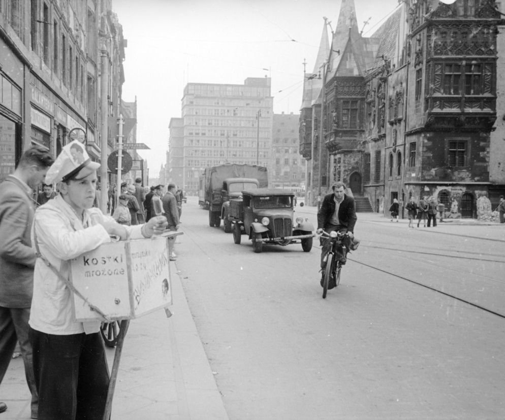 Czarno-białe zdjęcie ulicy we Wrocławiu z chłopcem sprzedającym lody z pojemnika, mężczyzną na rowerze, ciężarówkami oraz przechodniami. W tle widoczny jest wieżowiec Miejskiej Kasy Oszczędności, symbolizujący modernizację miasta, o czym możesz przeczytać na Architektura Murator Plus.