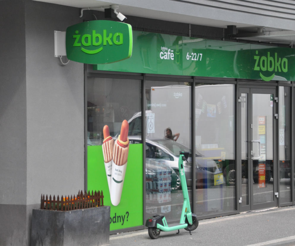 Witryna sklepu Żabka z plakatem hot dogów i hulajnogą przed wejściem. O handlu 15 sierpnia przeczytasz na Eska.