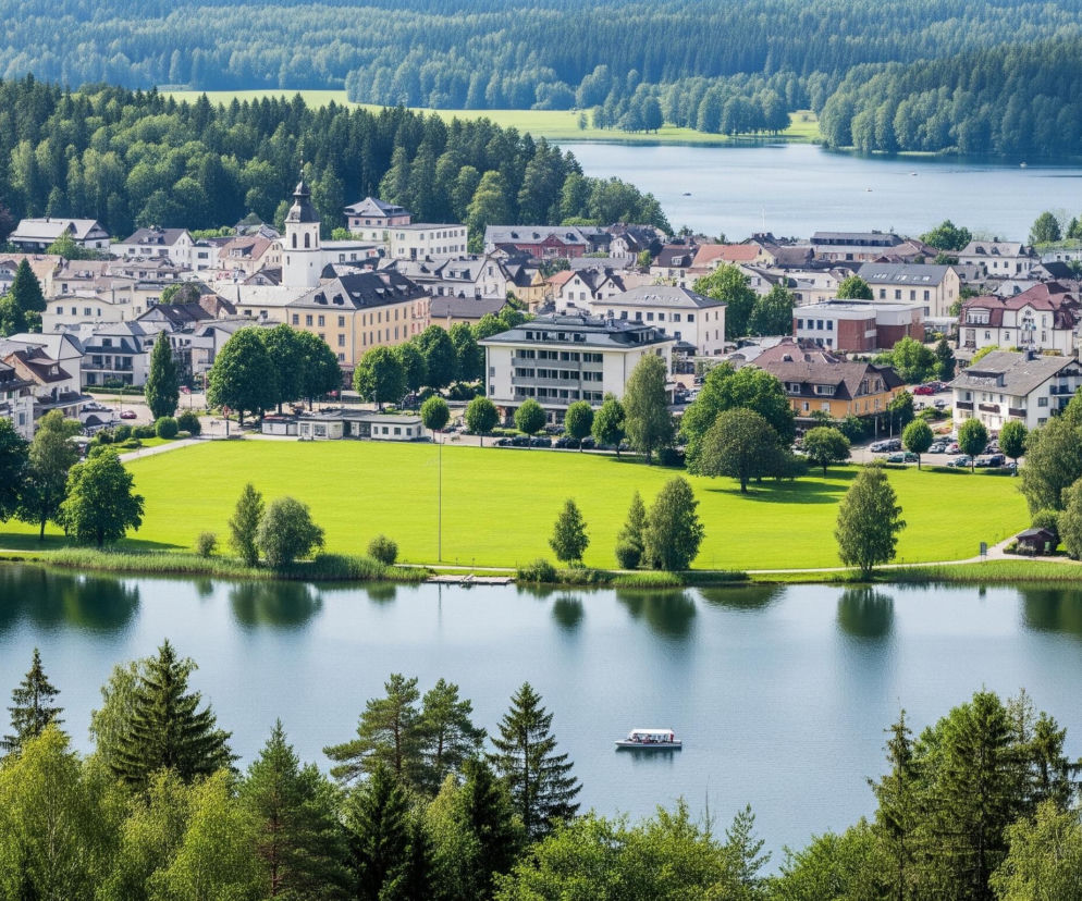 Duży, jasnoniebieski zbiornik wodny rozciąga się przez centrum kadru, z ciemnozielonym lasem otaczającym jego odległe brzegi. Na prawym brzegu, w oddali, widoczny jest długi, biały budynek, a obok niego pomarańczowy dach. Na powierzchni wody unosi się pojedyncza, biała łódź z pasażerami, widoczna w środkowej części obrazu. Po lewej stronie zbiornika wodnego, tuż za nim, rozciąga się małe miasteczko z licznymi budynkami o jasnych fasadach i spadzistych, ciemnych dachach, wśród których wyróżnia się biała wieża kościoła z ostrym szczytem.