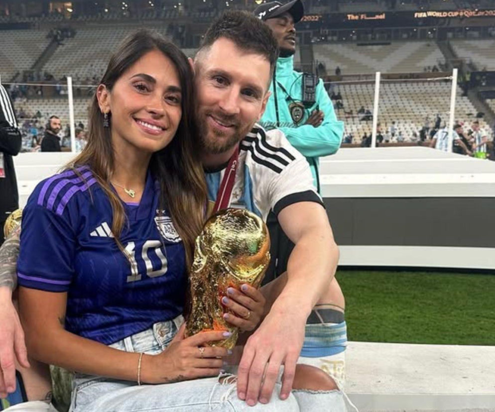 Antonela Roccuzzo i Leo Messi uśmiechają się do zdjęcia, trzymając trofeum, celebrując historyczny sukces Messiego na Mistrzostwach Świata. Antonela ma na sobie fioletową koszulkę Argentyny z numerem 10, a Leo biało-błękitną. Więcej o ich emocjach na naszym portalu.