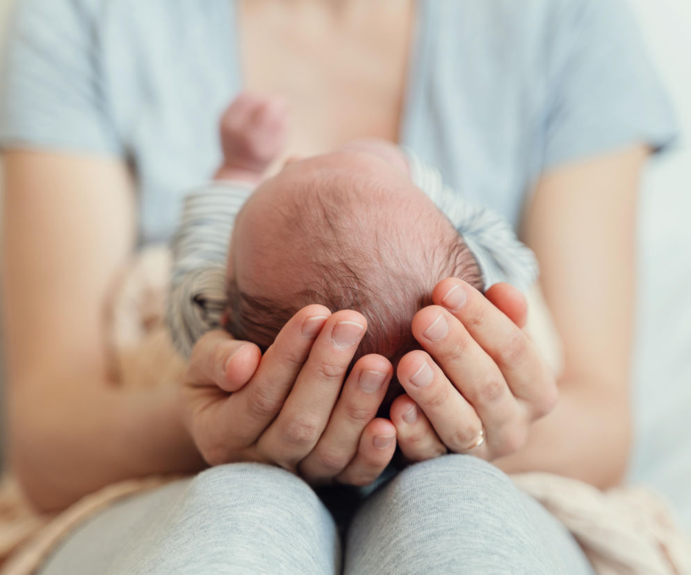 Dłonie osoby podtrzymują głowę niemowlęcia. O poszukiwaniu rodziny zastępczej dla chłopca przeczytasz na SE.