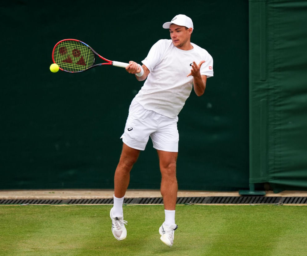 Polski tenisista Kamil Majchrzak w białym stroju podczas meczu na Wimbledonie, odbijający żółtą piłkę rakietą z pomarańczowo-czarną ramą, z zieloną ścianą w tle. Więcej o jego słodkiej zemście na trawie Wimbledonu przeczytasz na naszym portalu.