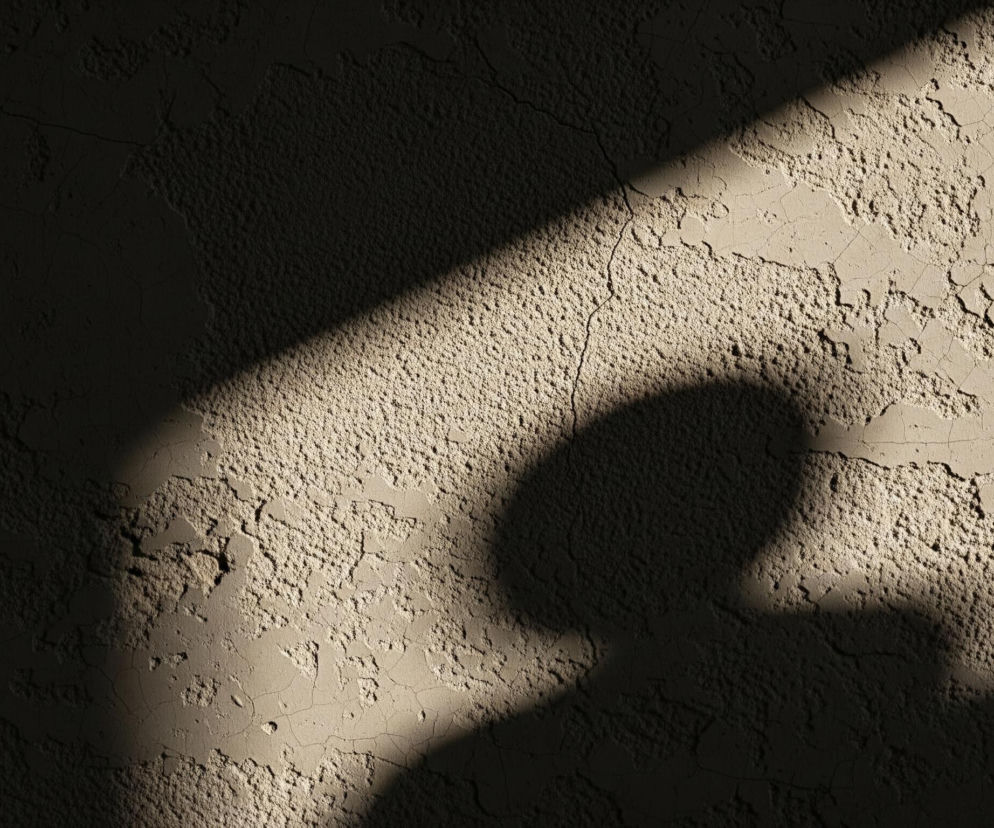 Teksturowana, chropowata ściana w odcieniach beżu i brązu jest oświetlona ukośnym pasem światła, tworzącym wyraźny kontrast między jasnymi i ciemnymi obszarami. Na jasnym, prawym fragmencie ściany widoczne są pęknięcia i nierówności tynku. Duży, owalny cień z lekko zaokrąglonym dnem, przypominający zarys głowy i ramion, rzuca się na środku ściany, częściowo wpadając w strefę światła i cienia, oraz przecinając ukośny pas światła.