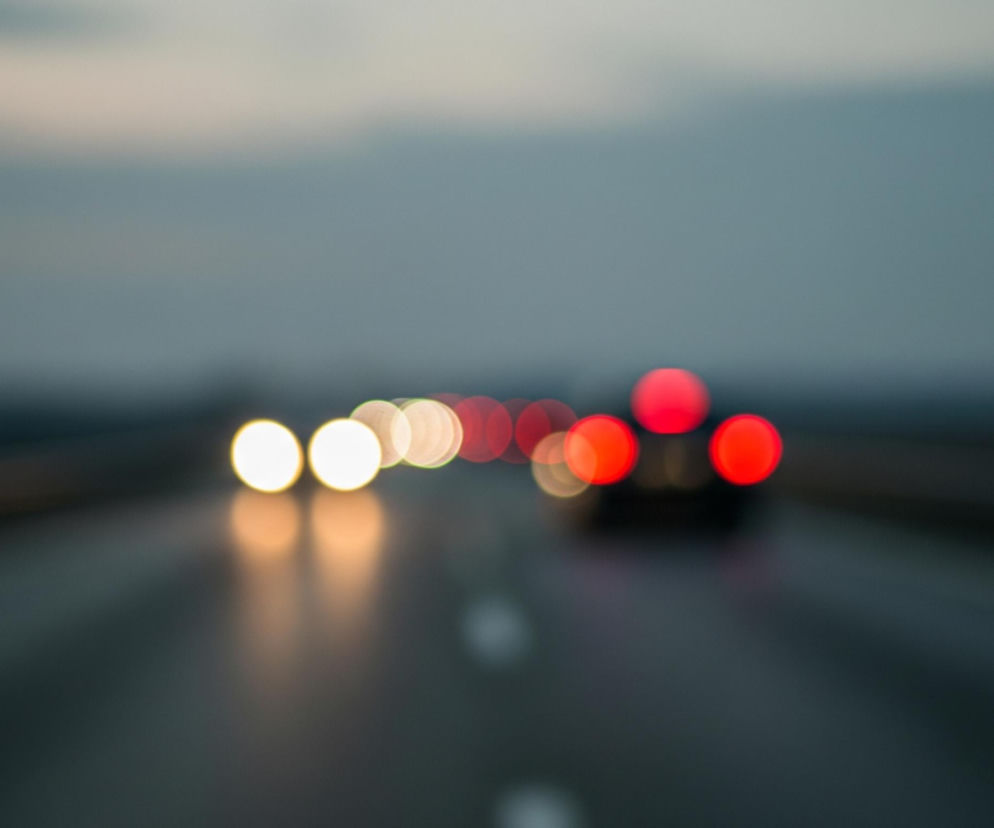 Rozmyte światła samochodów rozciągają się w głąb ciemnego, asfaltowego pasa drogi. Po lewej stronie widać szereg jasnych, ciepłych świateł, głównie żółtych i białych okręgów, z których niektóre odbijają się na wilgotnej nawierzchni. Po prawej, dalej na drodze, dominują czerwone, okrągłe światła tylne, również rozmyte. Tło stanowi zamglone, ciemnoszare niebo, jaśniejsze u góry, które płynnie przechodzi w ciemniejszy odcień horyzontu.