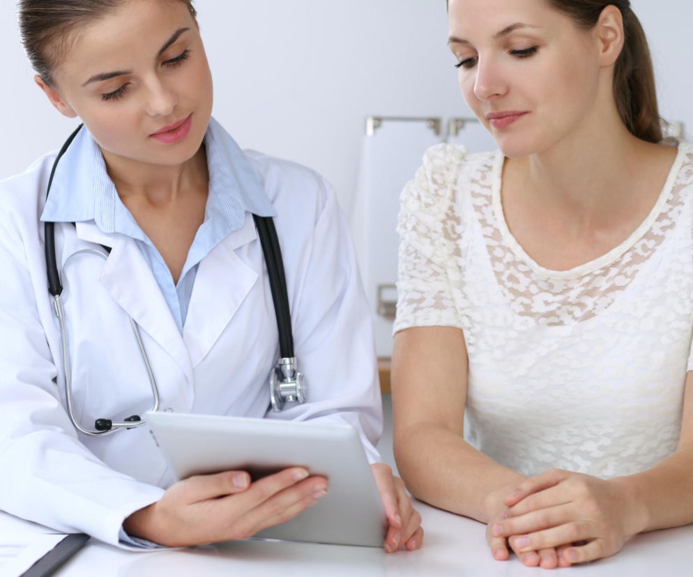 Lekarka w białym kitlu ze stetoskopem na szyi, prezentująca wyniki badań na tablecie pacjentce w białej, ażurowej bluzce. Temat badań profilaktycznych dla kobiet, o których więcej przeczytasz na portalu Poradnik Zdrowie.