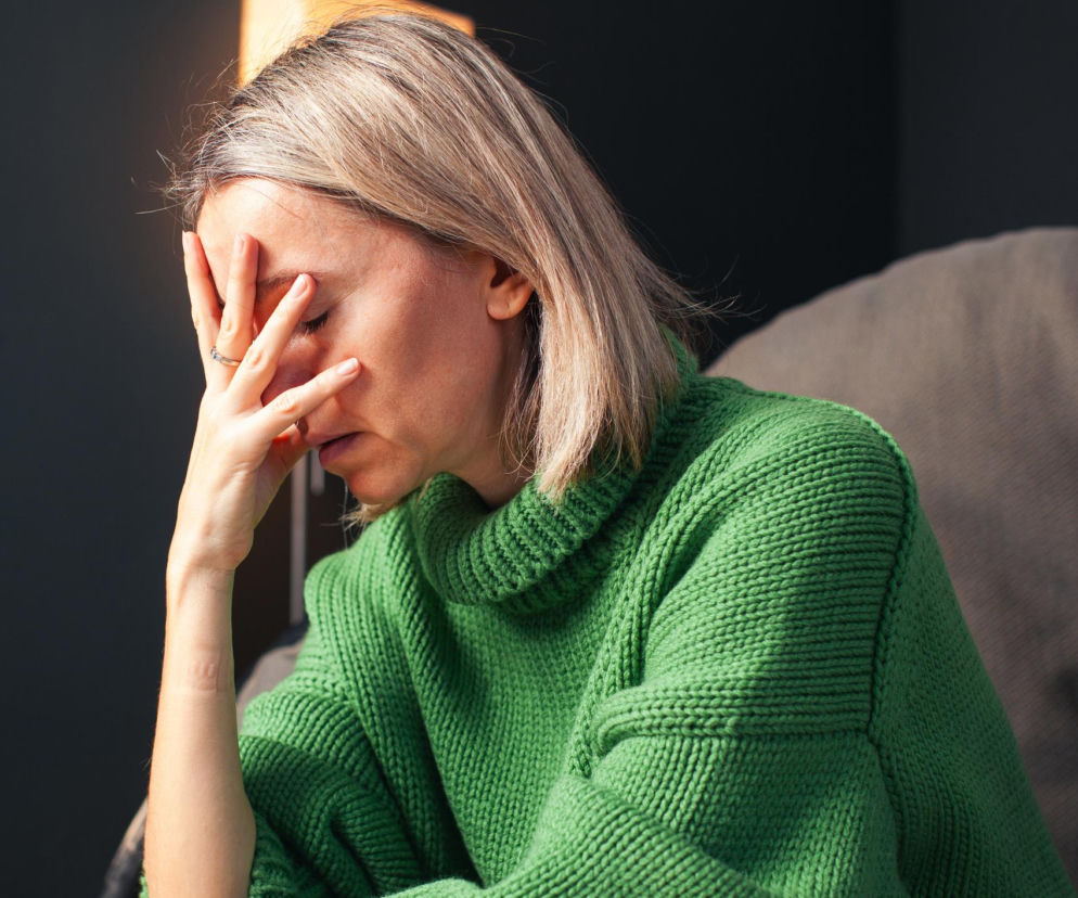 Zasmucona, przygnębiona kobieta, prawdopodobnie cierpiąca na głód skóry, z dłonią zasłaniającą twarz, ubrana w zielony, wełniany sweter. Poradnik Zdrowie wyjaśnia, jak brak dotyku wpływa na zdrowie psychiczne.