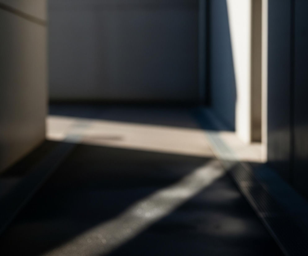 Przedstawiony jest tunel lub korytarz z wyraźnymi kontrastami światła i cienia, z silnym rozmyciem tła. Po lewej stronie dominuje ciemna ściana, natomiast w oddali, po prawej stronie, widać jasne, pionowe pasmo światła, prawdopodobnie pochodzące z zewnątrz. Podłoga korytarza jest częściowo oświetlona ukośnym pasmem słońca, tworzącym jaśniejszy obszar na ciemnej powierzchni. Na podłodze widoczne są również ciemne, równoległe elementy przypominające kratownicę lub kanalizację.