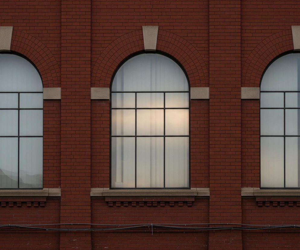 Czerwona ceglana ściana z trzema identycznymi oknami łukowymi, każde z nich posiada czarną ramę i jest podzielone na sześć prostokątnych sekcji u dołu oraz jedną dużą półkolistą sekcję u góry. Nad każdym oknem łuk z cegieł ozdobiony jest klinem z jasnego kamienia, a pod nim prosty parapet z tego samego materiału. W środkowym oknie widoczne jest intensywne, jasne światło, podczas gdy pozostałe dwa odbijają zróżnicowane, stonowane odcienie szarości i błękitu, sugerując zasłony lub żaluzje. Poniżej okien biegnie szereg poziomych kabli.