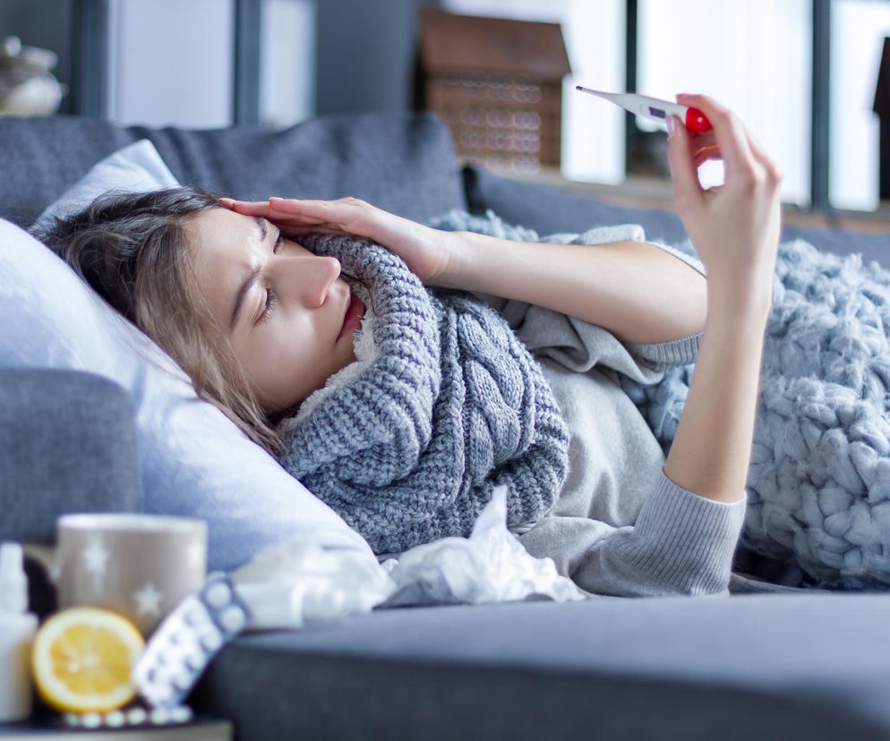 Młoda kobieta w szarym swetrze i szaliku leży na kanapie pod kocem, trzymając termometr i dotykając czoła, co symbolizuje objawy grypy i gorączkę. Obok leżą chusteczki, cytryna, tabletki i spray na gardło, wskazujące na leczenie w domu. Więcej o grypie na Poradnik Zdrowie.