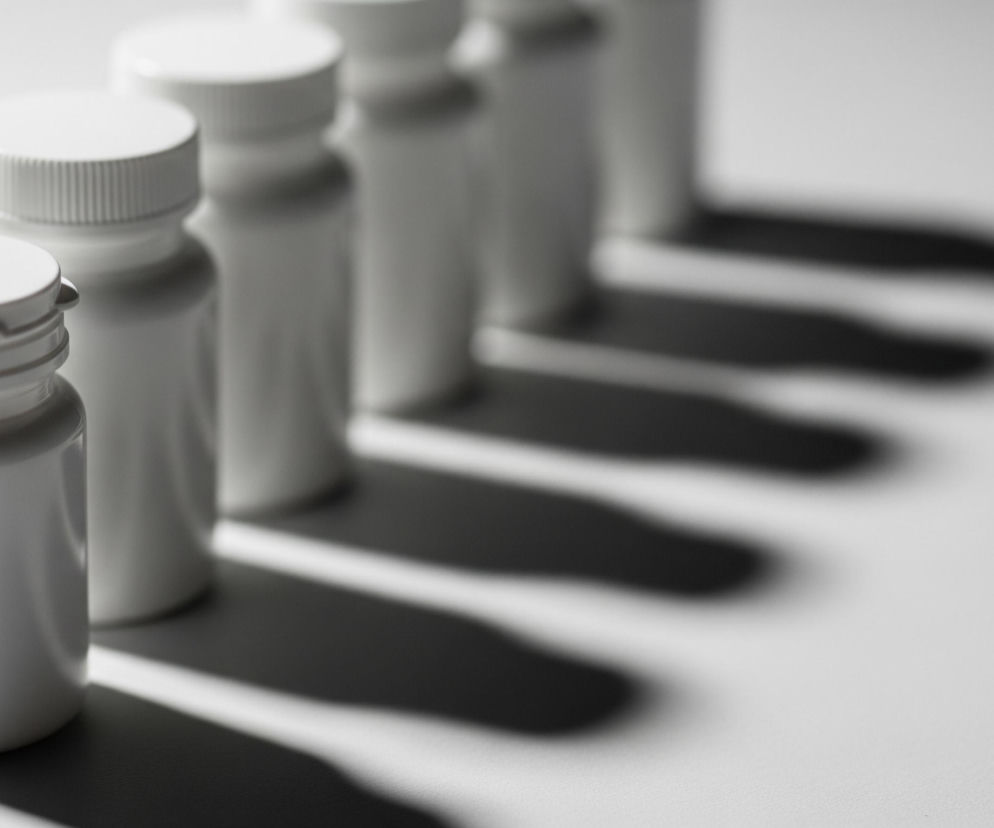 Rząd białych, walcowatych pojemników z nakrętkami ustawiony jest na jasnej powierzchni, biegnąc w głąb obrazu od lewej do prawej, zbiegając się. Pierwszy pojemnik na pierwszym planie jest ostry, a kolejne stają się coraz bardziej rozmyte. Od każdego pojemnika pada długi, ciemny cień w prawo, tworząc równoległe smugi cienia na jasnym tle.