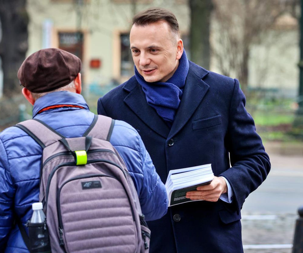 Łukasz Gibała w ciemnym płaszczu rozmawia z przechodniem, trzymając plik dokumentów. O wyborach w Krakowie przeczytasz na Eska.