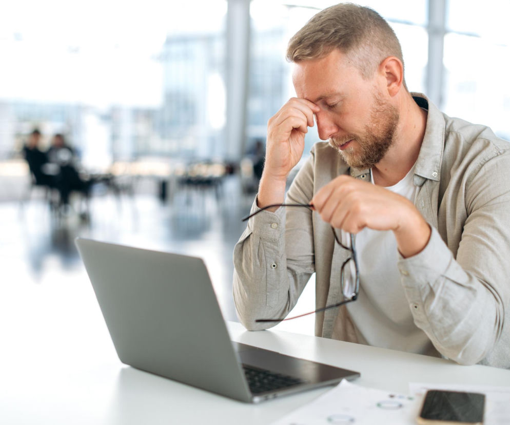 Mężczyzna z brodą w koszuli, z okularami w ręku, masuje skroń, siedząc przy biurku z laptopem w biurze. Prawdopodobnie odczuwa ból głowy. Zgodnie z artykułem na Poradnik Zdrowie, ból głowy w pracy często wynika z odwodnienia lub wahań cukru.