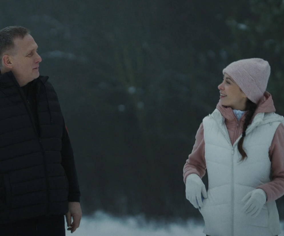 Artur (Robert Moskwa) i Joanna (Dominika Sakowicz) podczas potajemnego spotkania w lesie, oboje ubrani w zimowe kurtki i czapki, stoją na śniegu, patrząc na siebie z uśmiechem. Mężczyzna jest w czarnej puchowej kamizelce, a kobieta w białej. Więcej o schadzce Artura z Joanną w lesie przeczytasz na Super Seriale.