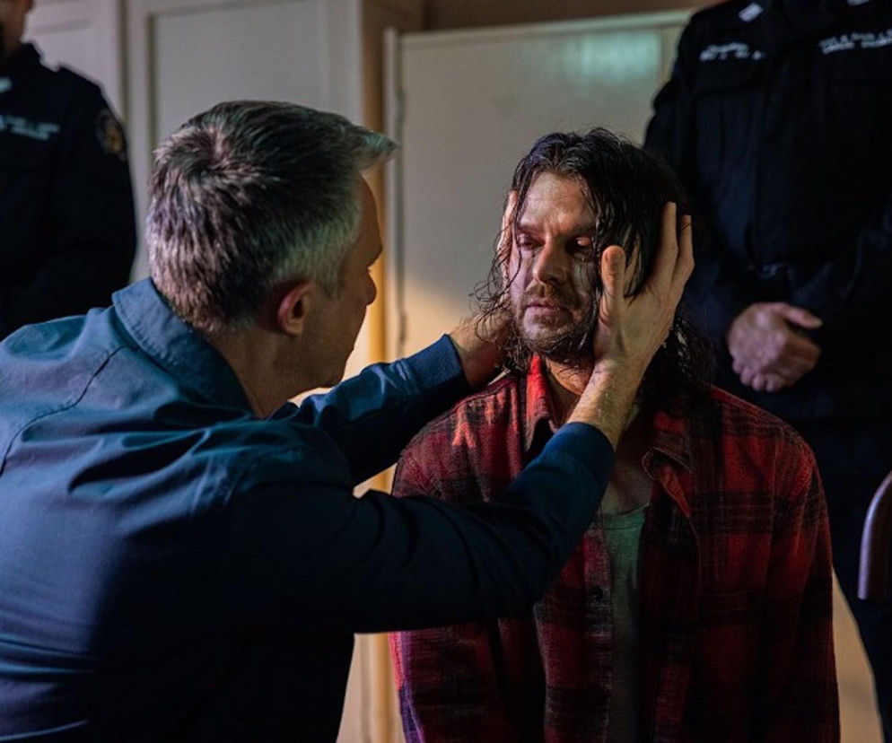 Artur (Łukasz Płoszajski) z serialu Pierwsza miłość opiera dłonie na twarzy skatowanego i wycieńczonego Pawła Krzyżanowskiego (Mateusz Kościukiewicz), który ma zmierzwione włosy i krwawy ślad pod okiem. W tle, niewyraźni strażnicy więzienni. Więcej o dramatycznych losach bohaterów na Super Seriale.