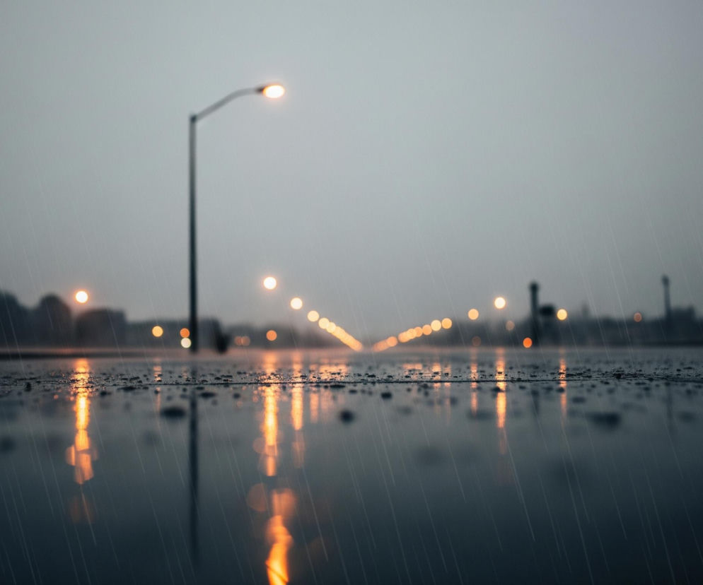 Mokry asfalt lub inna utwardzona powierzchnia zajmuje dolną połowę obrazu, odbijając ciepłe, pomarańczowe światła uliczne. Na pierwszym planie widoczne są pojedyncze krople deszczu, a liczne smugi sugerują padający deszcz. W centrum, w tle, wyróżnia się pojedyncza lampa uliczna z jasnym, pomarańczowym światłem, a dalej w oddali rozciąga się linia podobnych, nieostrych świateł, tworząc perspektywę drogi. Niebo jest pochmurne i szare, harmonizując z ciemnymi, rozmytymi sylwetkami budynków w tle.