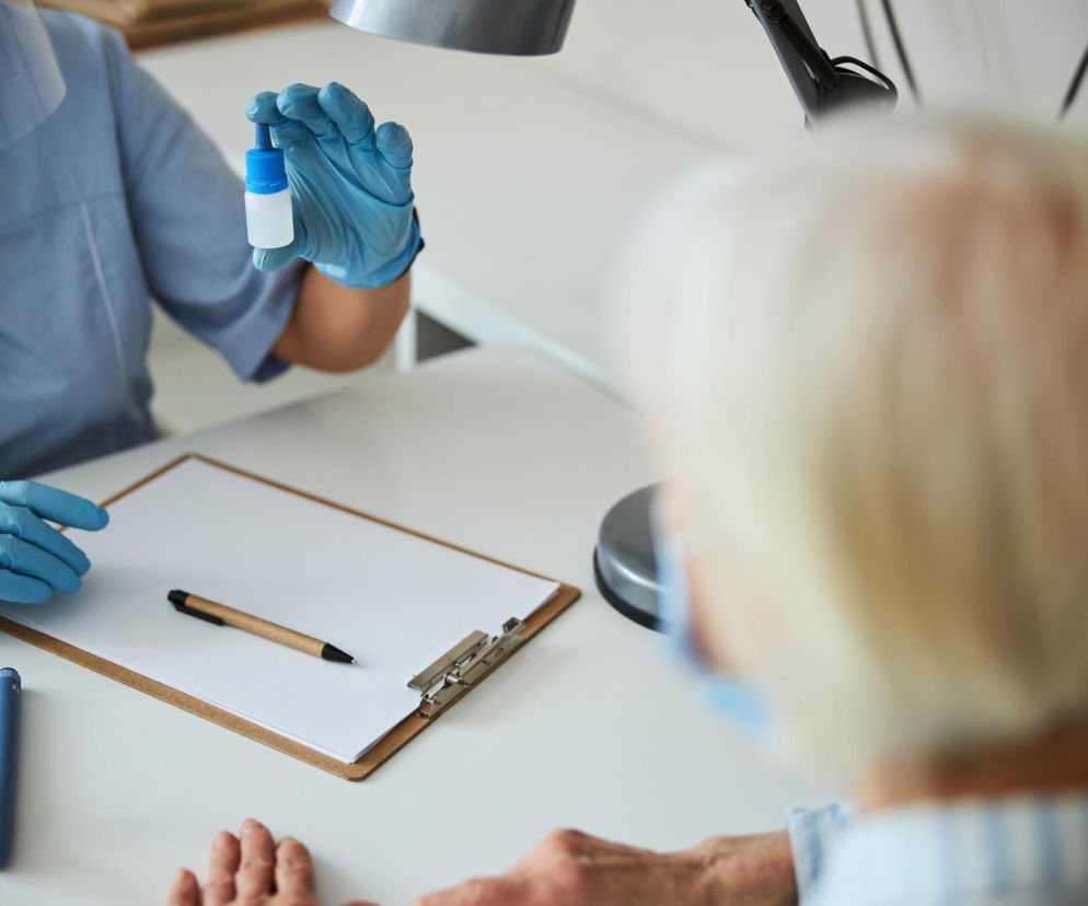 Pracowniczka medyczna w niebieskich rękawiczkach i osłonie twarzy trzyma małą buteleczkę z lekarstwem, prezentując ją starszemu pacjentowi. Przed nią na biurku leży schowek z pustą kartką, długopis, inhalator do nosa. Zdjęcie ilustruje temat ukrytych skutków infekcji wirusowych i jest częścią artykułu na Poradnik Zdrowie.