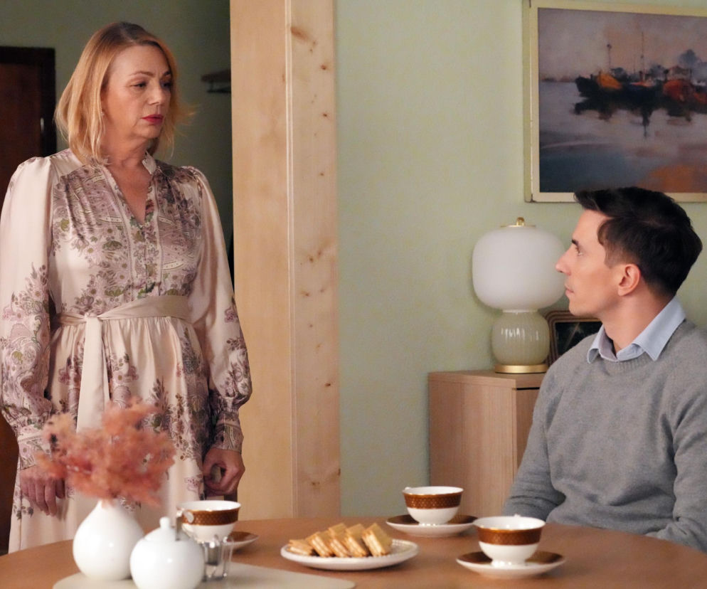 Aktor Jakub Sokołowski jako Tomek siedzi przy stole z filiżankami i ciasteczkami, spoglądając w prawo, podczas gdy aktorka Maja Barełkowska jako Wanda stoi obok, patrząc z zamyśleniem. Ten moment z serialu Barwy szczęścia i inne Super Seriale znajdziesz w naszym serwisie.